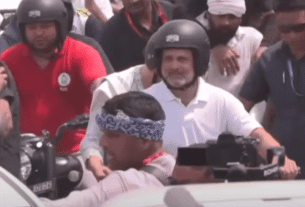 A group of people, some wearing helmets, navigate through a crowd with motorcycles on a busy street during the daytime.