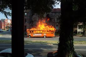 A fire burns on the side of a bus, with flames and smoke visible against the vehicle's exterior.