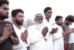 A group of men stands in a line with their hands clasped in prayer. They wear white clothing, and the background is a soft, overcast sky, creating a serene atmosphere.