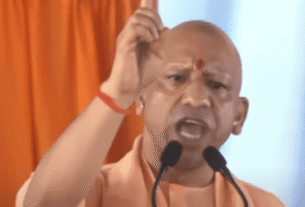 A person in orange attire passionately speaks at a podium, gesturing upwards with one hand. The backdrop features an orange curtain, conveying intensity.