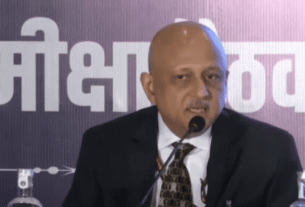 A man in a suit and tie speaks into a microphone at a conference. The backdrop displays Hindi text. The setting is formal and professional.
