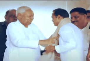 A group of men in traditional white clothing smile as one man drapes a shawl over another, conveying warmth and camaraderie in a ceremonial setting.