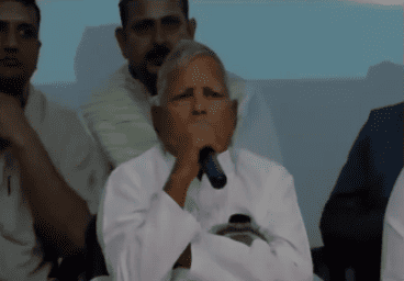 An elderly man in white traditional attire speaks into a microphone at a public event, surrounded by attentive seated audience members.