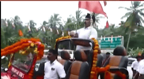 A man in a white shirt and black cap stands in an open jeep decorated with flowers and flags. Palm trees and a road sign are visible in the background.