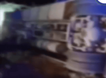 Blurred image of a large vehicle turned on its side, possibly a truck or bus, on a dimly lit street at night, suggesting an accident scene.