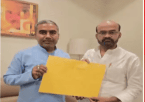 Two men stand side by side in a room, holding a yellow folder together. The man on the left wears a blue shirt, and the one on the right is in white. There's a painting and a lamp in the background, suggesting an office setting.